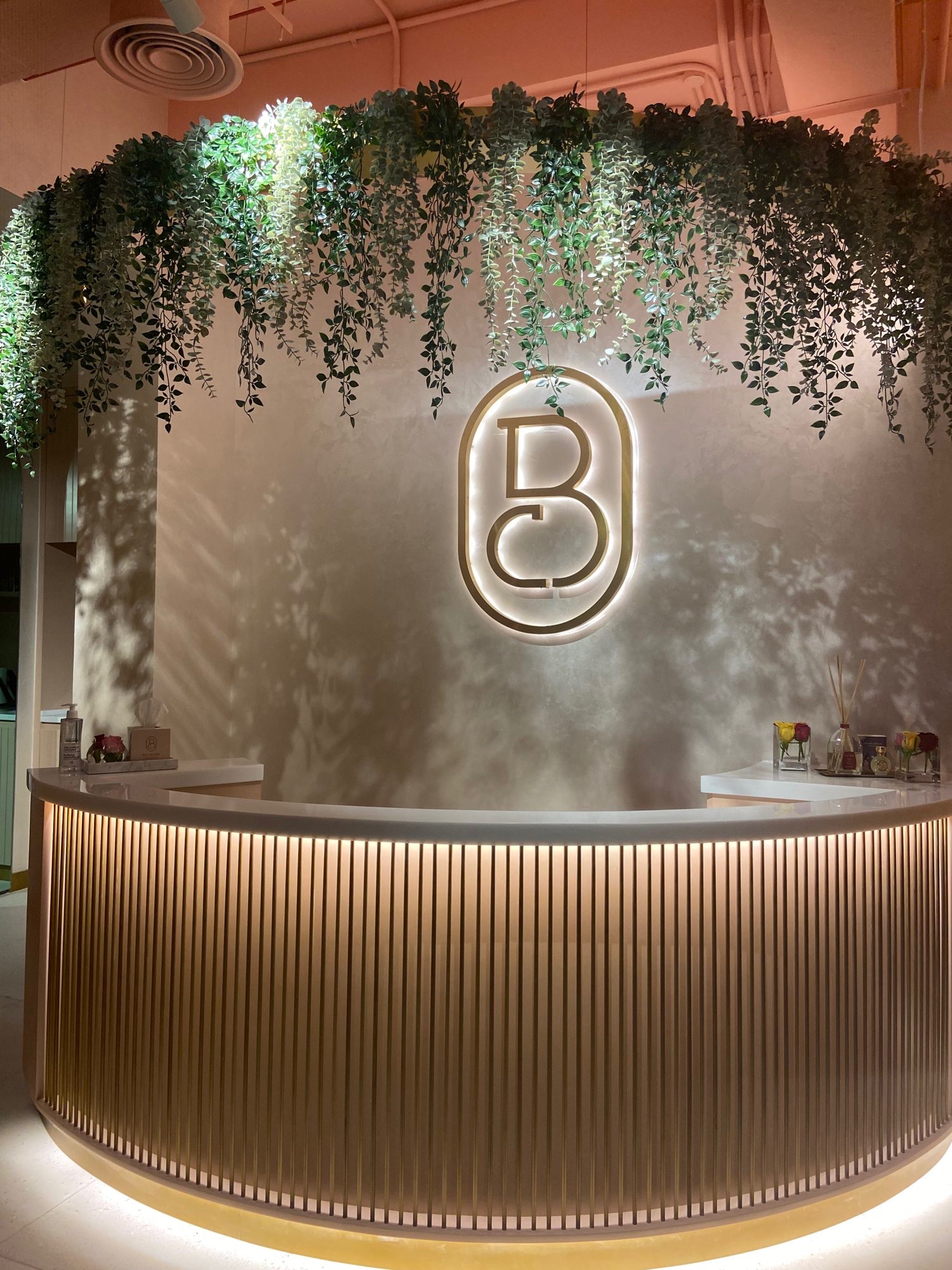 The reception at Bellacure Al Mamoura: a curved gold desk under trailing plants, with the Bellacure monogram lit on the wall