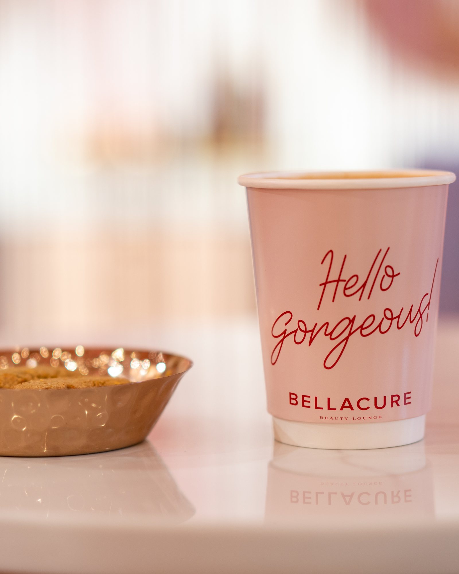A Hello Gorgeous Bellacure coffee cup beside a gold dish of cookies at the Al Seef cafe