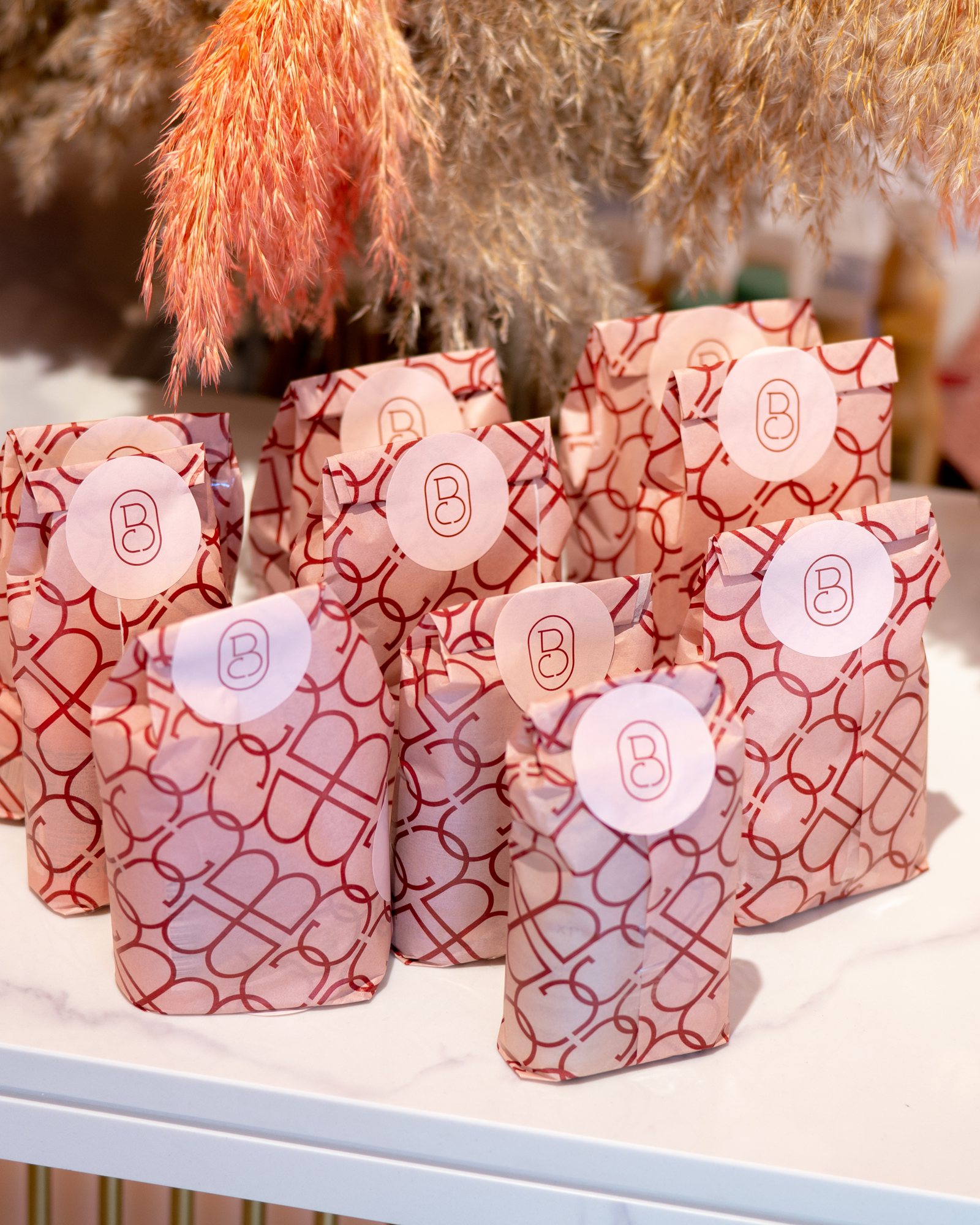Bellacure gift bags in the red monogram print, lined up on a marble counter under pampas grass