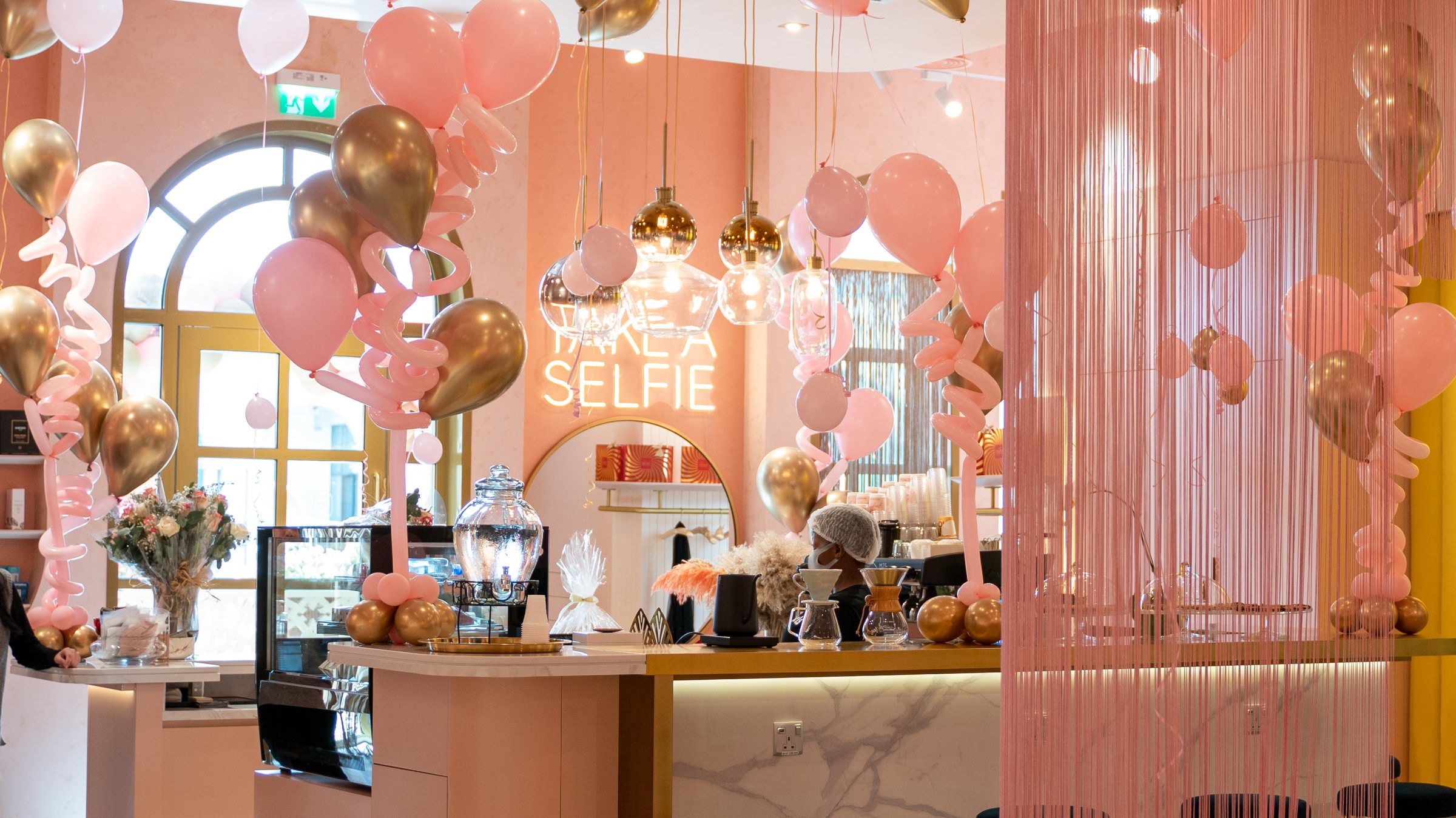 The Bellacure café at Al Seef filled with pink and gold balloons for a Mother's Day event, the Take a Selfie neon glowing behind the counter