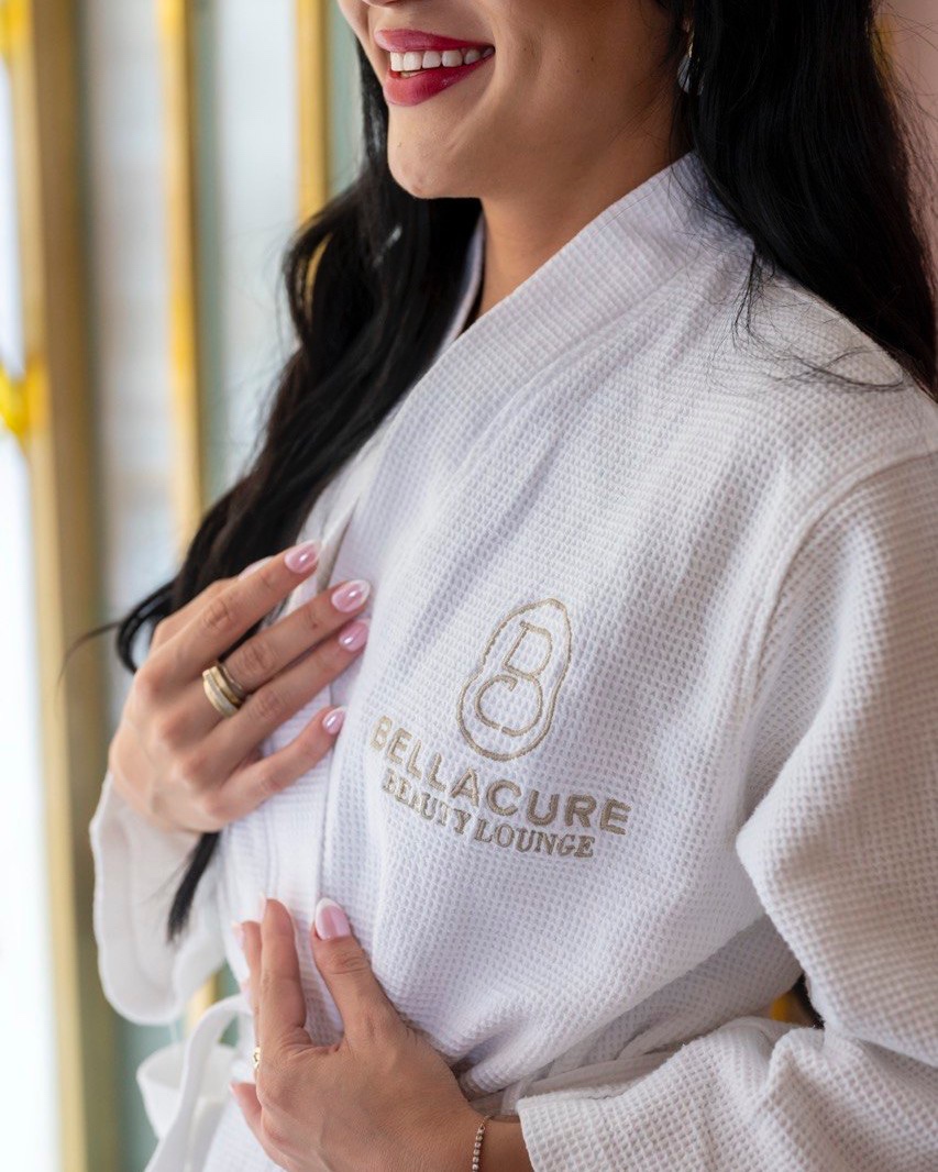 A smiling guest in a white Bellacure Beauty Lounge waffle robe with the gold monogram