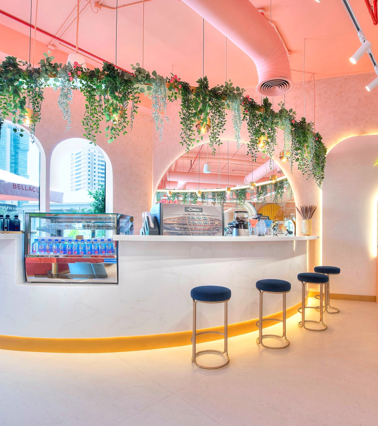 The Bellacure café at Sheikha Fatima Park: a curved marble counter with gold stools, a pastry case and trailing plants under the pink arches