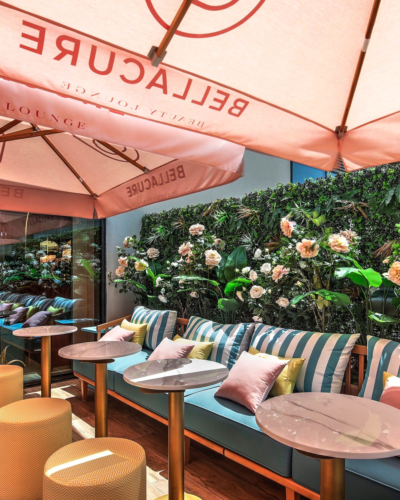 The café terrace at Bellacure Sheikha Fatima Park: striped sofas and marble tables against a wall of roses, under pink Bellacure umbrellas